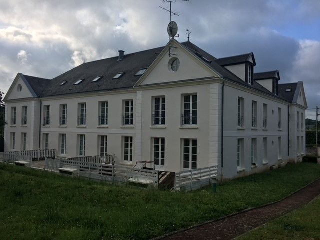 Bâtiment blanc de deux étages avec de nombreuses fenêtres, se détachant sur un ciel nuageux, avec de l'herbe au premier plan.