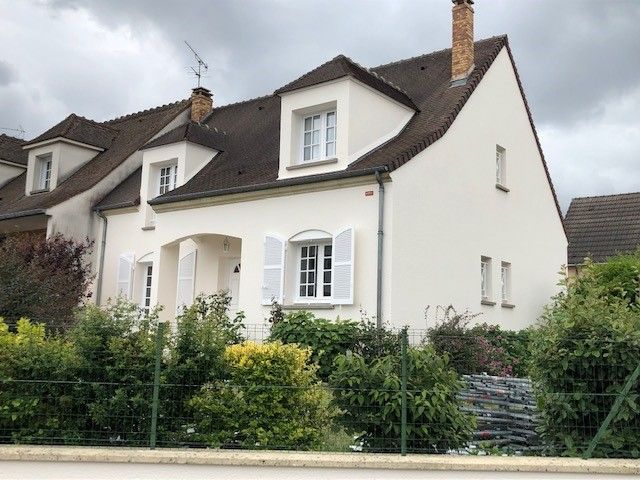 Maison blanche au toit marron, avec volets et lucarnes, derrière une clôture verte et des buissons.
