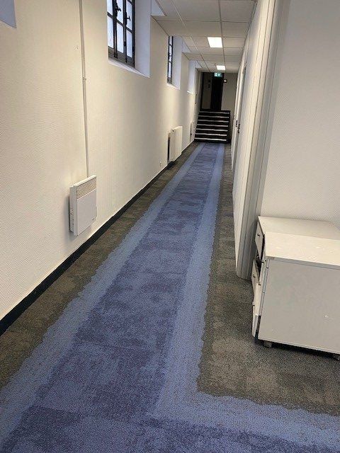 Long couloir avec un tapis bleu, des murs blancs et un escalier au loin.