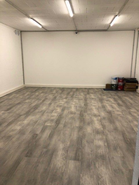 Pièce vide avec sol imitation bois et murs blancs. Des pots de peinture et du matériel sont posés dans un coin.