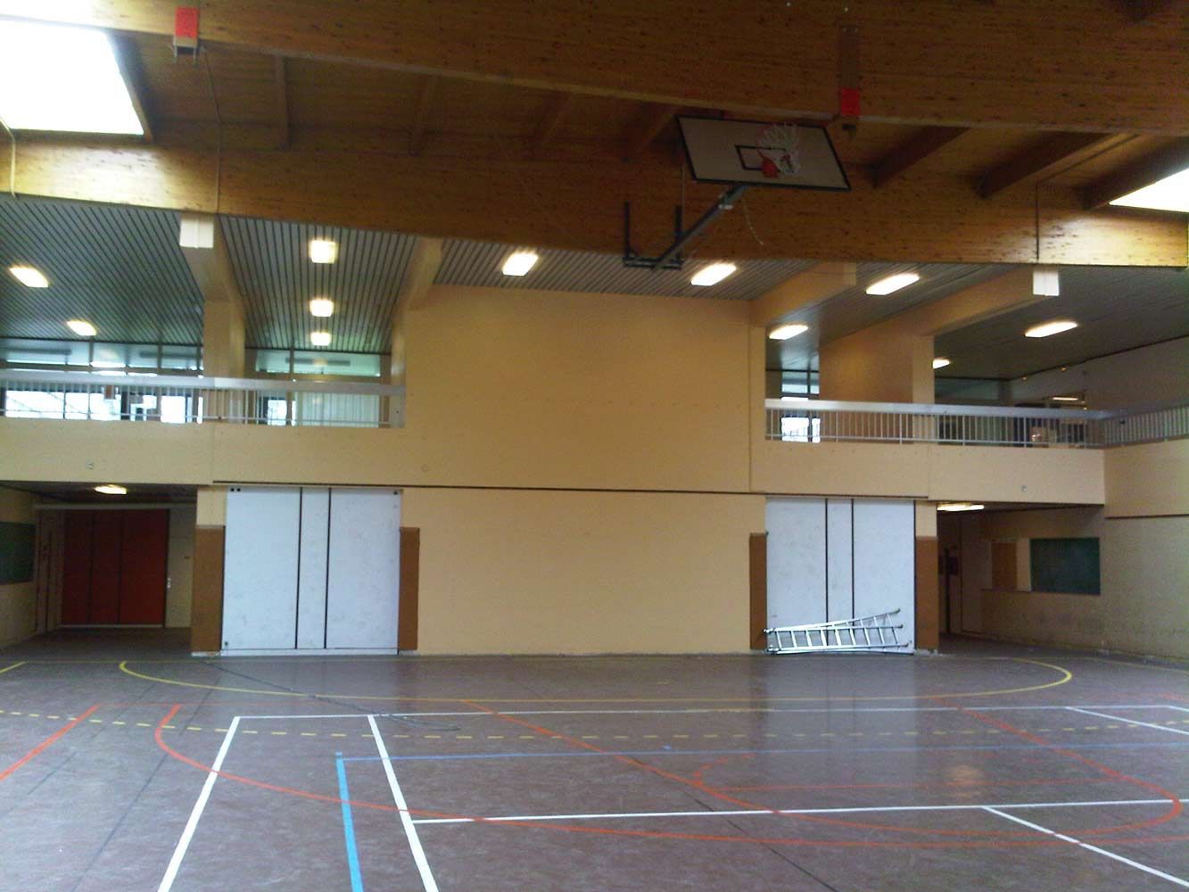 Gymnase vide avec panier de basket et murs et sol de couleur claire, bâtiment à deux étages.