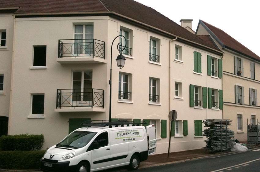 Immeuble beige de trois étages avec des volets verts, des balcons noirs et une camionnette blanche garée devant.