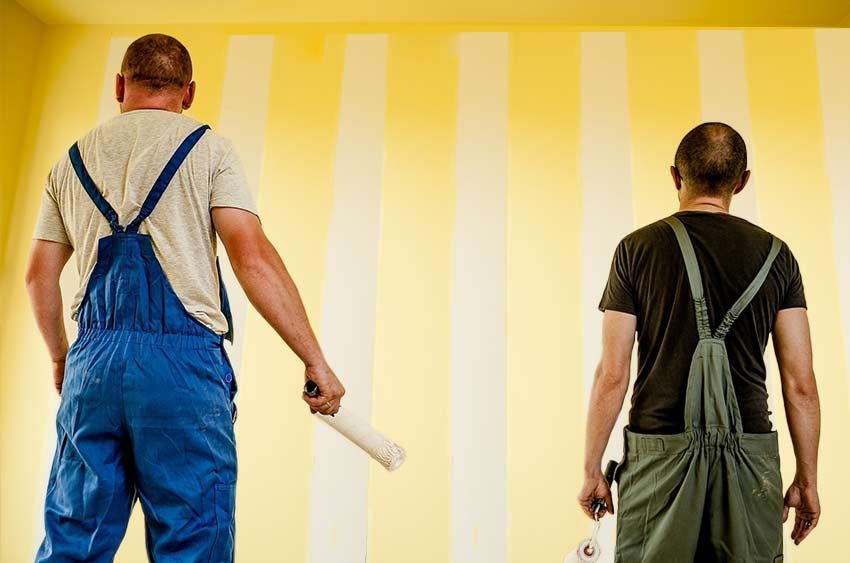 Deux ouvriers en salopette peignent des rayures jaunes sur un mur à l'aide de rouleaux à peinture.