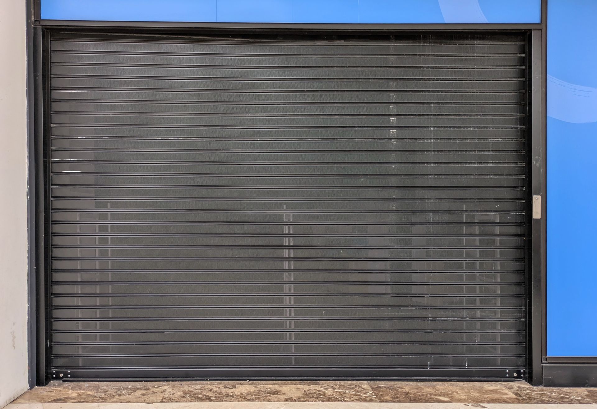 Un volet roulant de sécurité noir et horizontal abaissé sur une porte à côté d'un mur bleu.