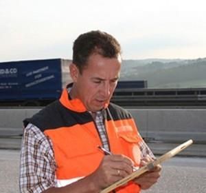 Ein Mann in einer orangefarbenen Weste schreibt auf einem Klemmbrett.