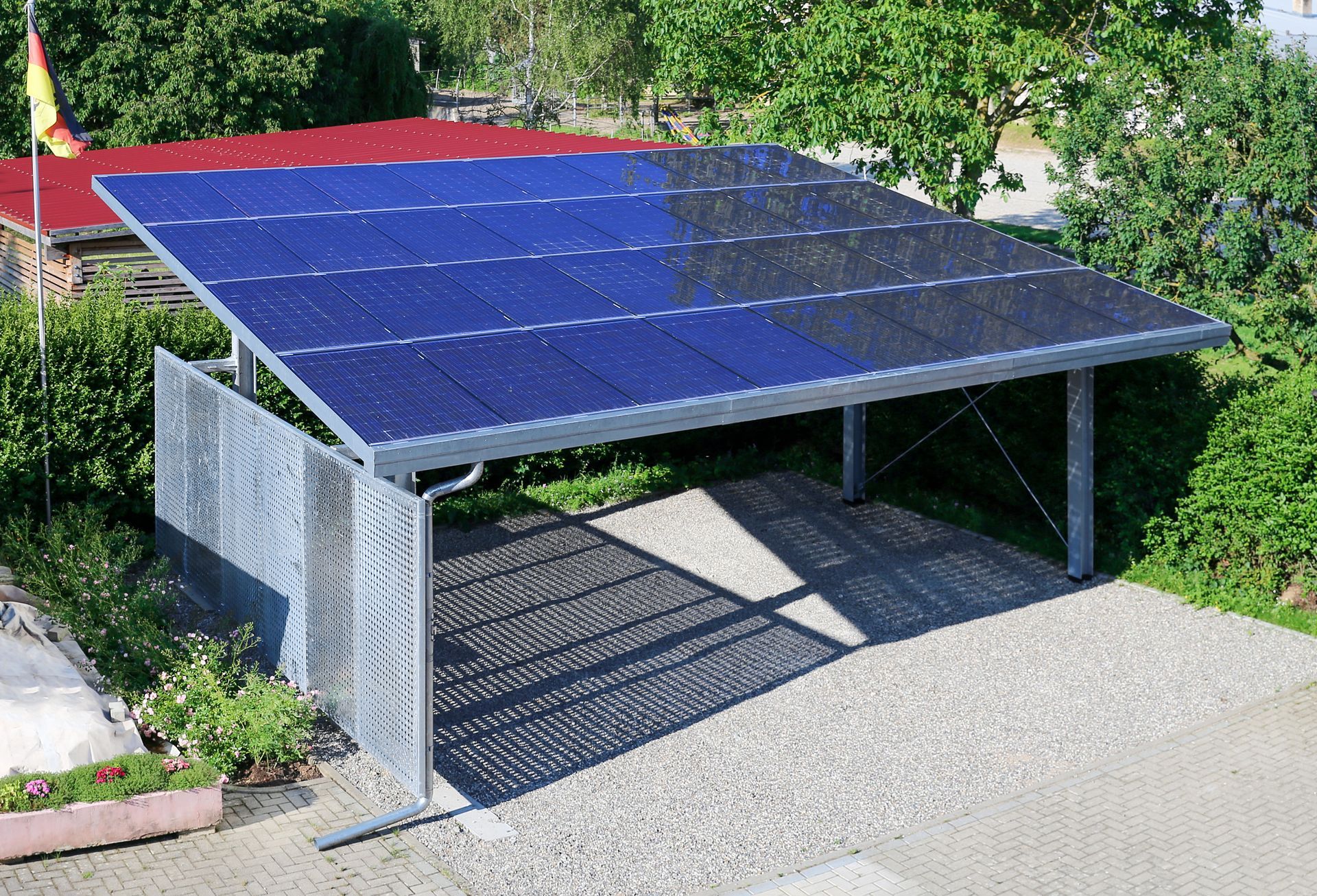 Abri voiture avec panneaux solaires bleus sur le toit, structure métallique, allée en gravier et végétation environnante.
