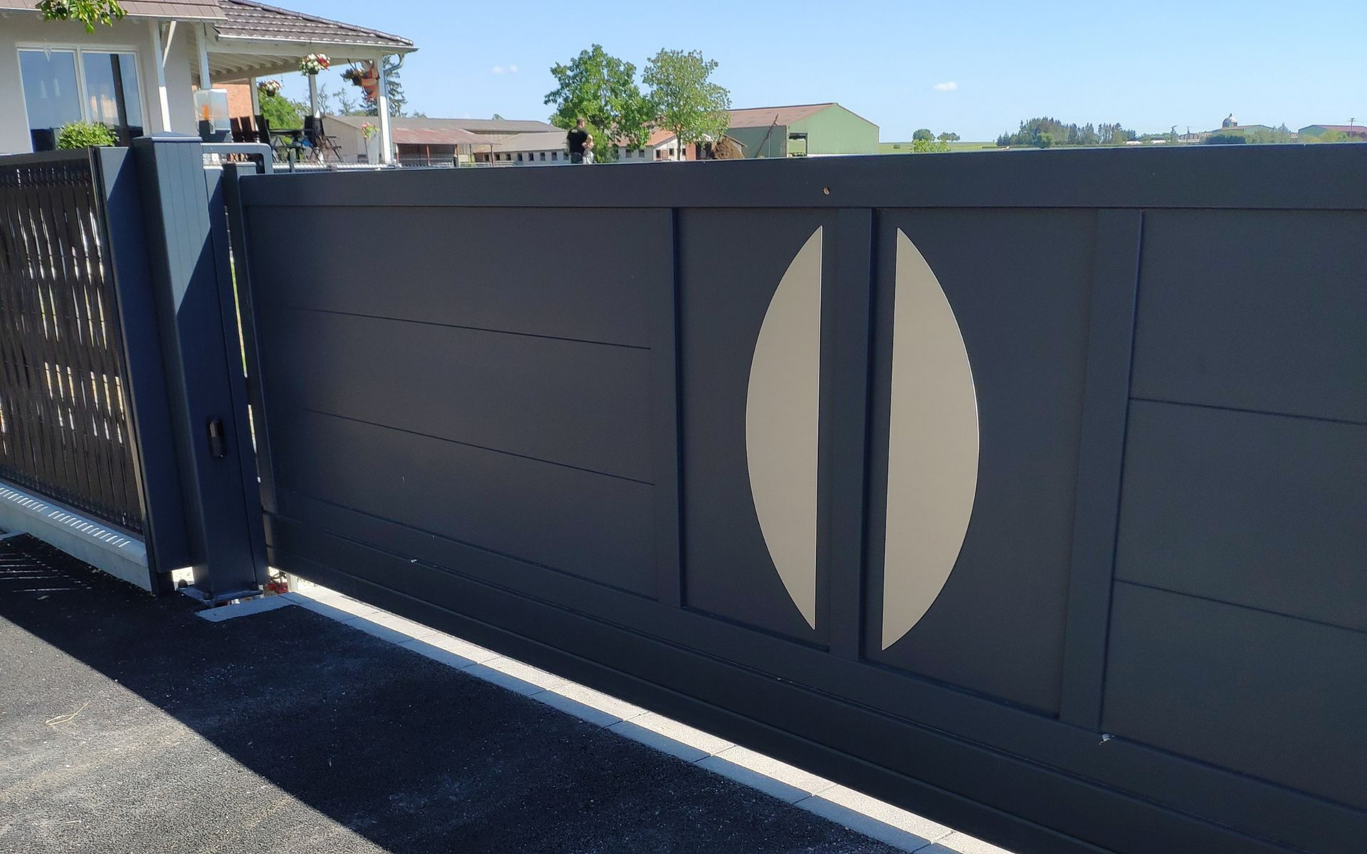 Portail coulissant gris foncé à motif ovale, à côté d'une clôture à motifs devant une maison.