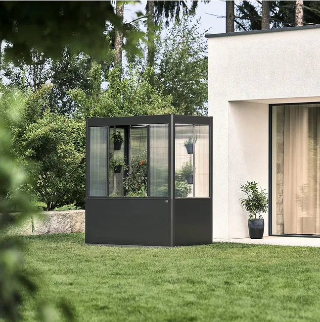 Serre extérieure gris foncé sur une pelouse, à côté d'un bâtiment blanc. Végétation à l'intérieur et autour de la serre.