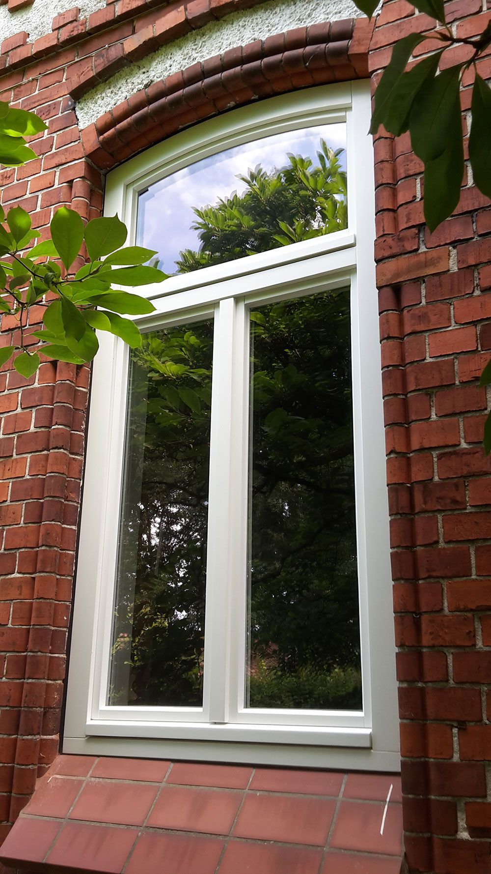 Ein weißes Fenster an einem roten Backsteingebäude, in dem sich Bäume spiegeln.