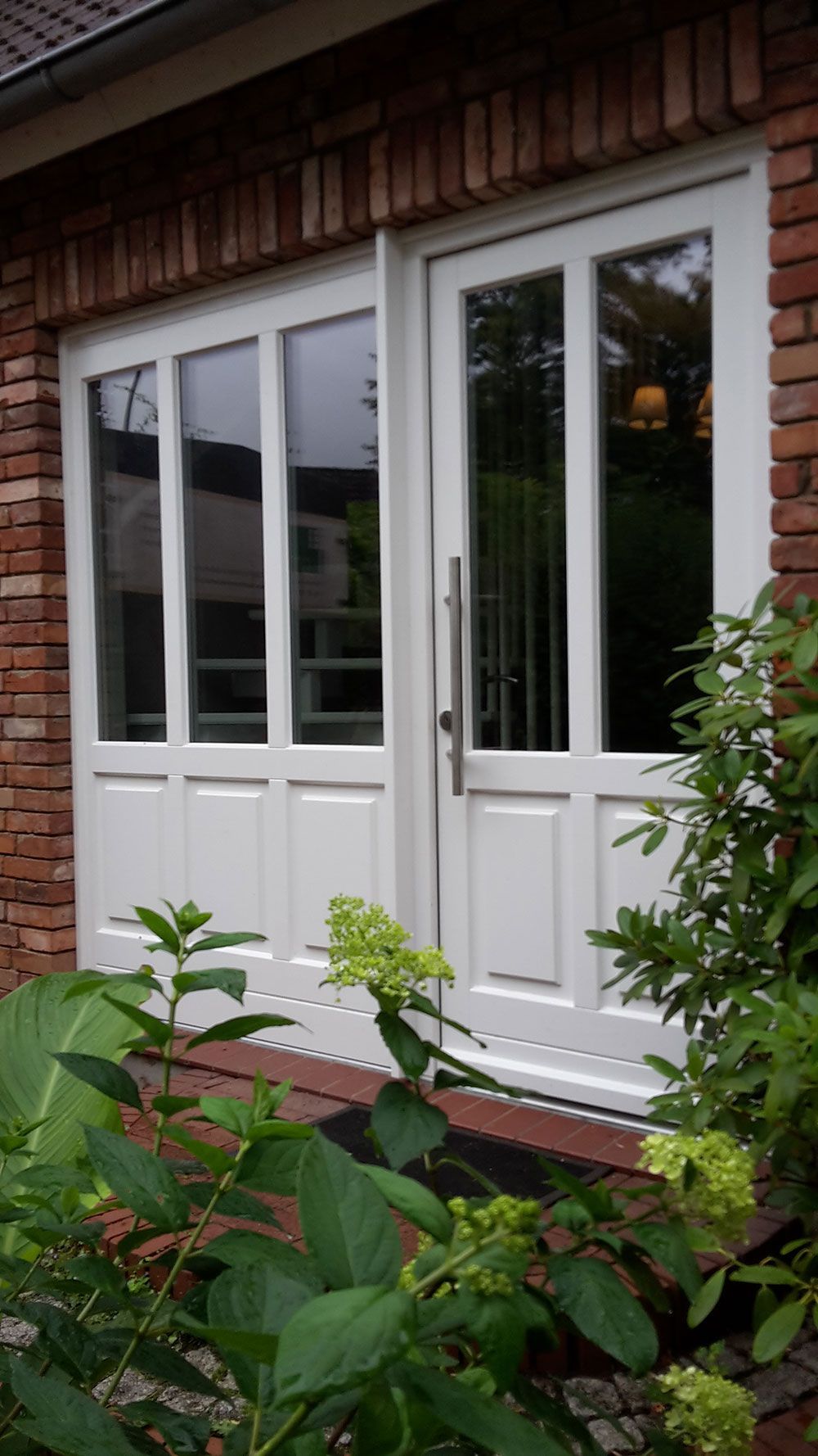 Eine weiße Tür mit einer Backsteinmauer im Hintergrund ist von Pflanzen umgeben.