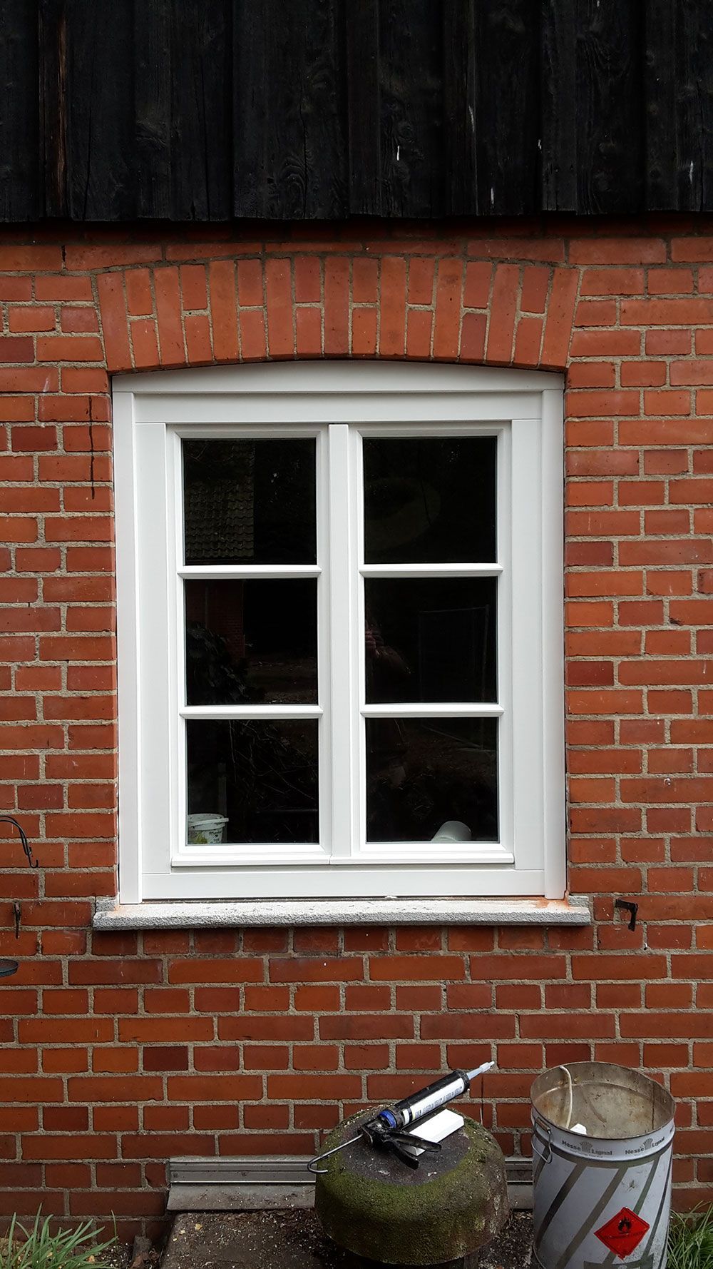 Neben einem Eimer Farbe befindet sich ein weißes Fenster an einer roten Backsteinwand.