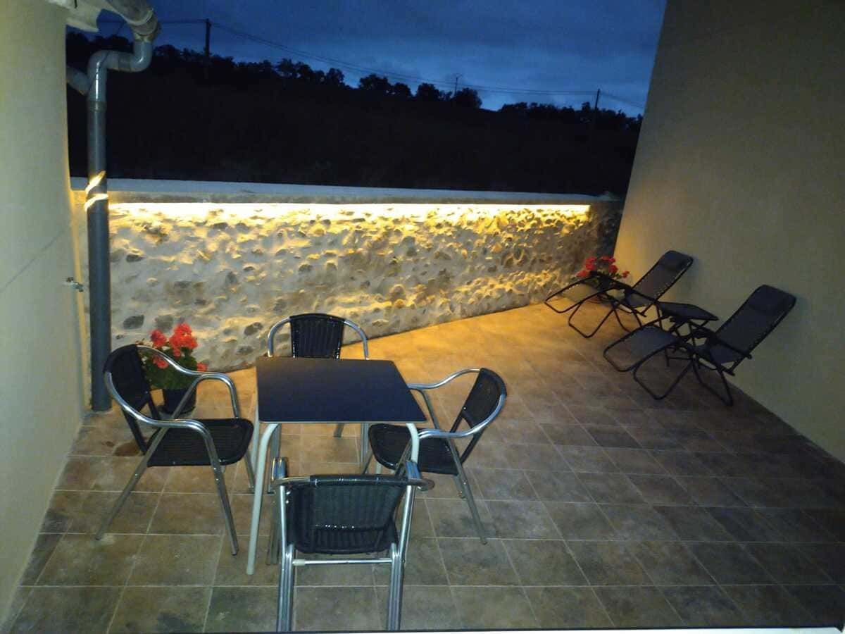 Casa de campo rústica de piedra con contraventanas verdes, plantas en macetas y vistas al campo bajo un tejado de tejas.