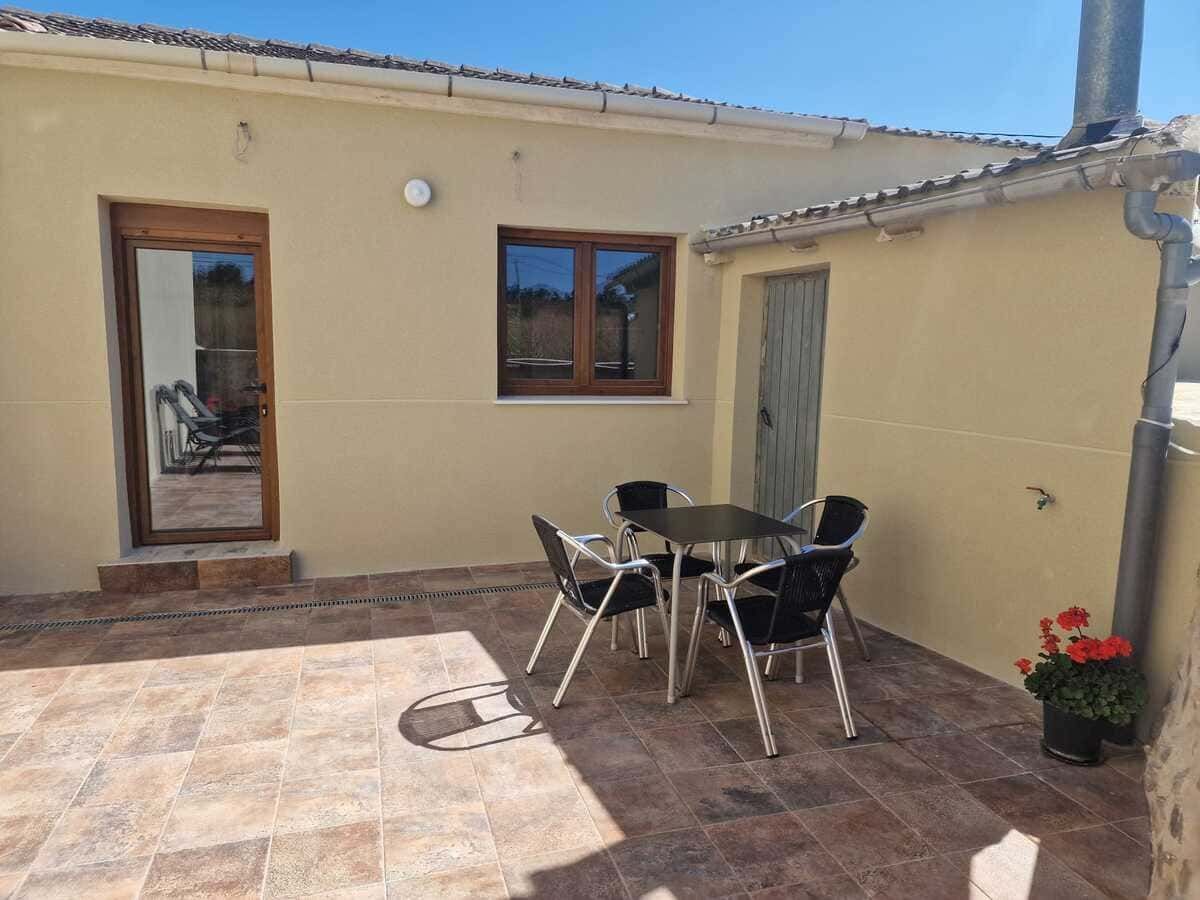 Chimenea exterior de piedra con tiro en un patio ajardinado, rodeada de plantas en macetas y una casa rústica.