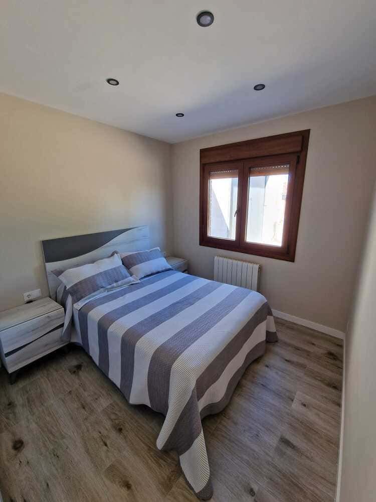 Dormitorio pequeño con una cama individual, ropa de cama a rayas, mesita de noche, ventana y suelo de aspecto madera.
