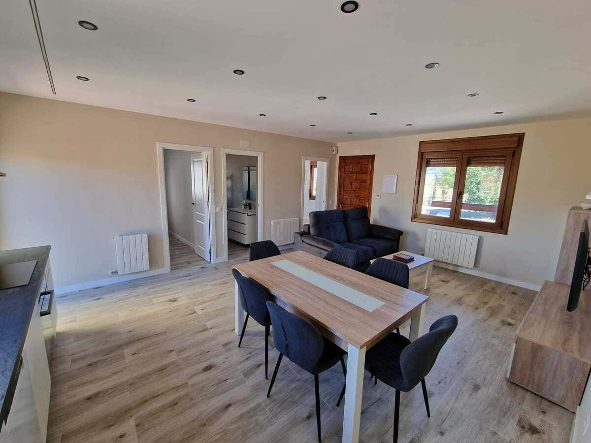 Salón luminoso y diáfano con mesa de comedor, sofá oscuro y grandes ventanales con persianas de madera.
