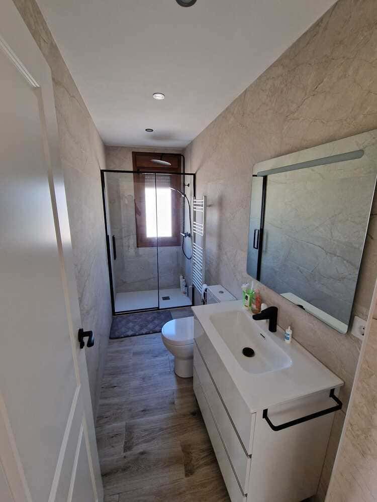 Baño moderno y estrecho con lavabo, inodoro, ducha y un espejo grande, iluminado por una ventana pequeña.