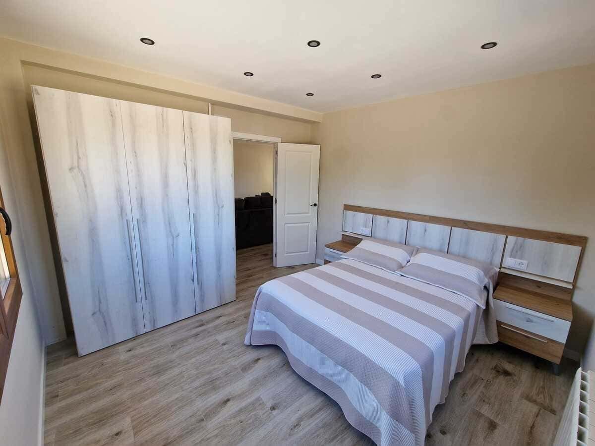 Dormitorio luminoso con una cama, un armario, mesitas de noche y suelo de madera clara.