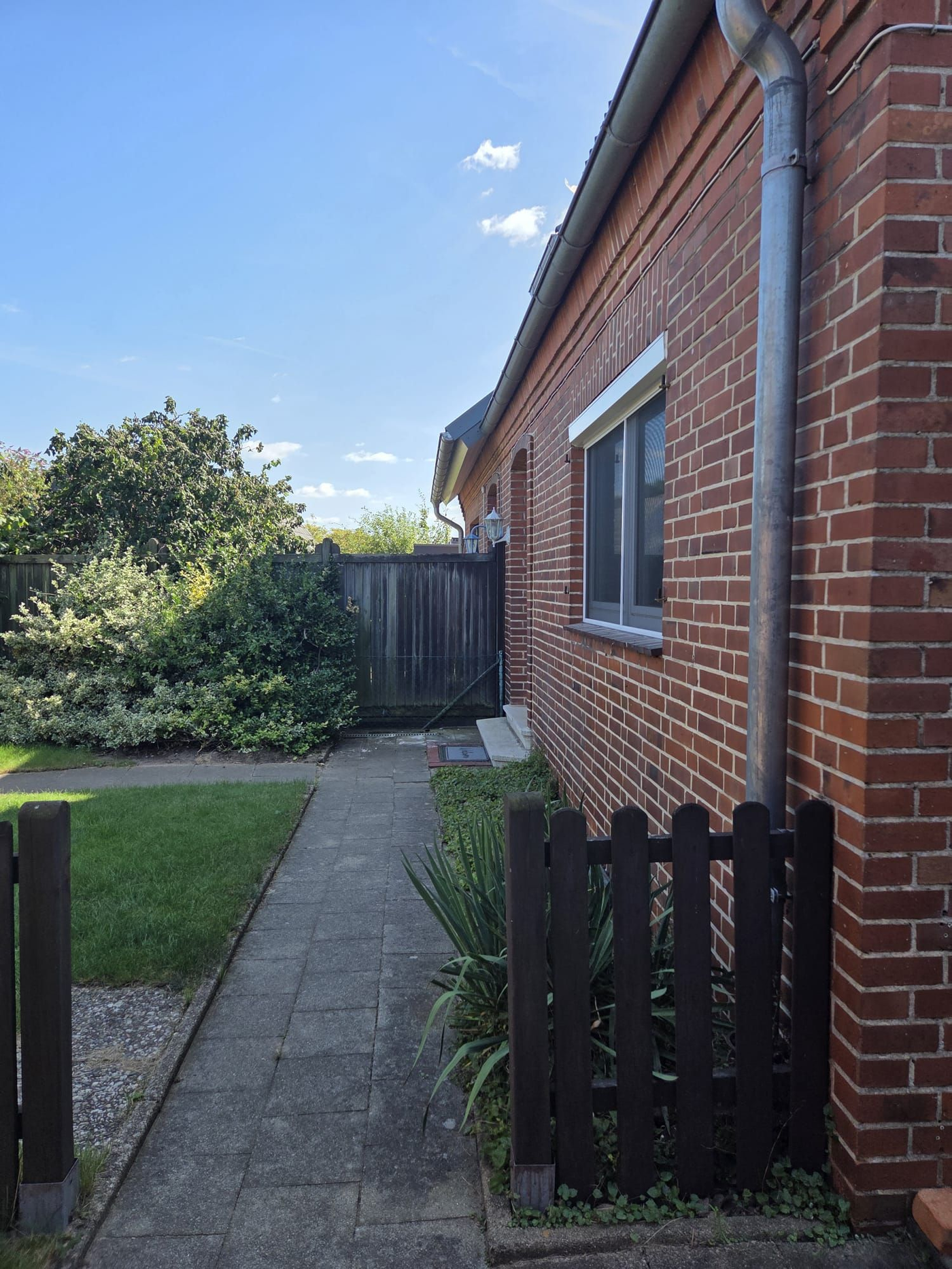 Backsteingebäude mit einem Weg, der zu einem Holztor führt, umgeben von einem Zaun und Grünflächen.