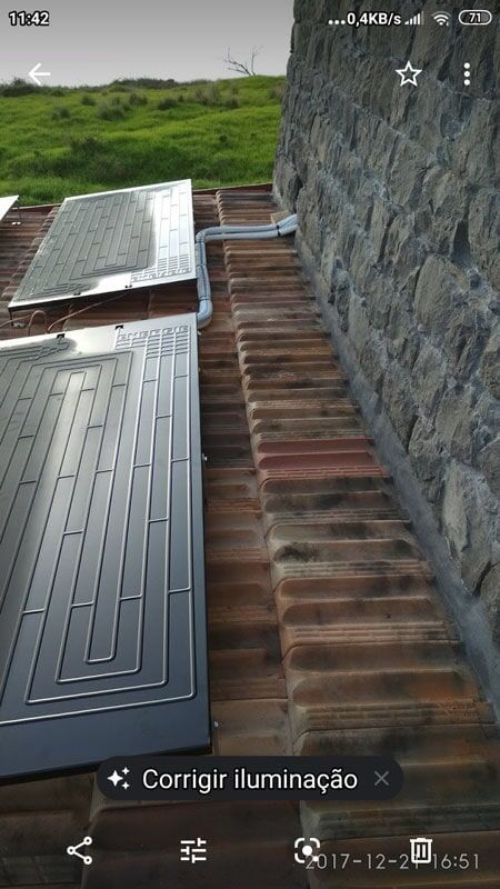 Painéis solares instalados em um telhado de telhas ao lado de um muro de pedra; vegetação ao fundo.