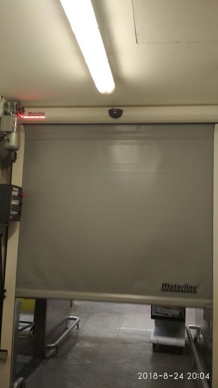 Porta industrial cinza de enrolar, parcialmente aberta. Luz laser vermelha acima, luz fluorescente no teto.