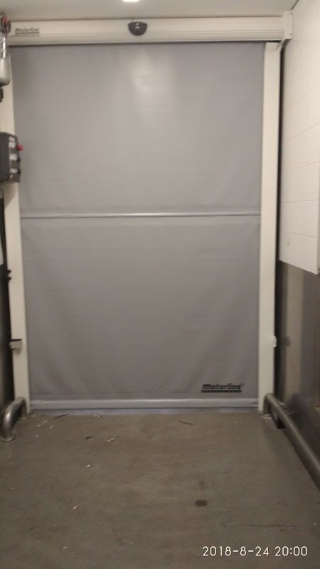 Porta de enrolar industrial cinza em moldura branca, com painel de controle à esquerda e câmera acima.