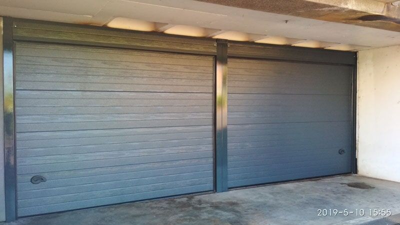 Duas portas de garagem cinzentas, lado a lado, sob uma cobertura de concreto.