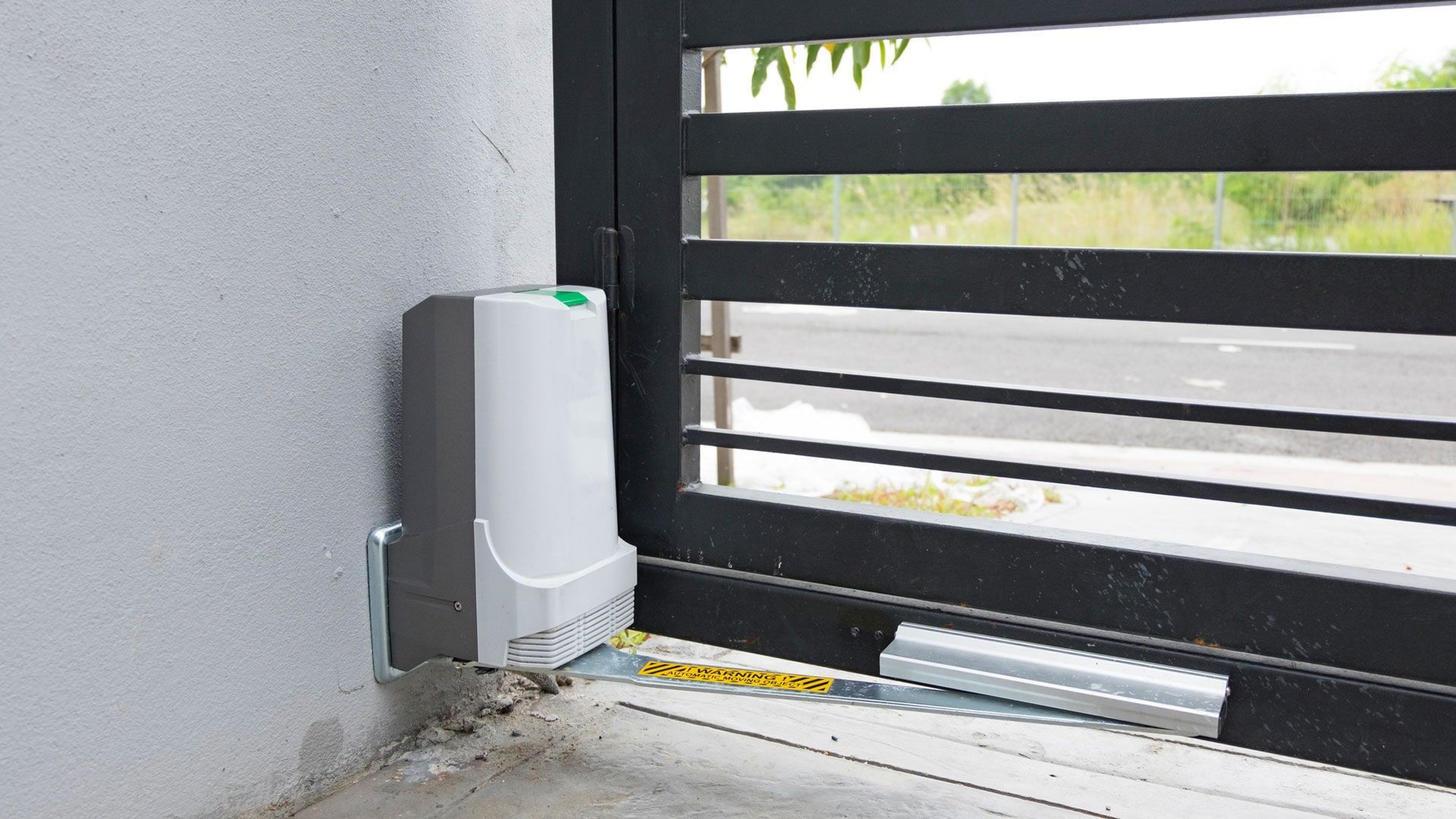 Abridor automático de portão montado em uma parede branca, conectado a um portão de metal preto.