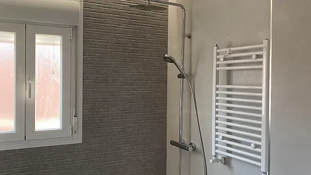Baño con ventana, pared de ducha de azulejos, cabezal de ducha y calentador de toallas blanco.