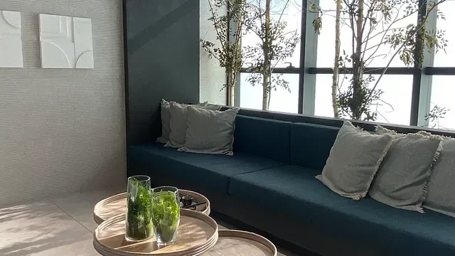 Un banco empotrado de color verde azulado con almohadas, pequeñas mesas redondas con jarrones y grandes ventanales con vista.