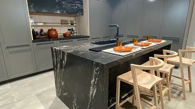 Cocina gris moderna con isla, encimera oscura, taburetes de madera, platos y gabinetes naranjas.