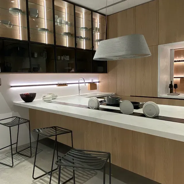 Cocina moderna con gabinetes de madera clara, encimeras blancas, gabinetes con frente de vidrio y asientos de barra.