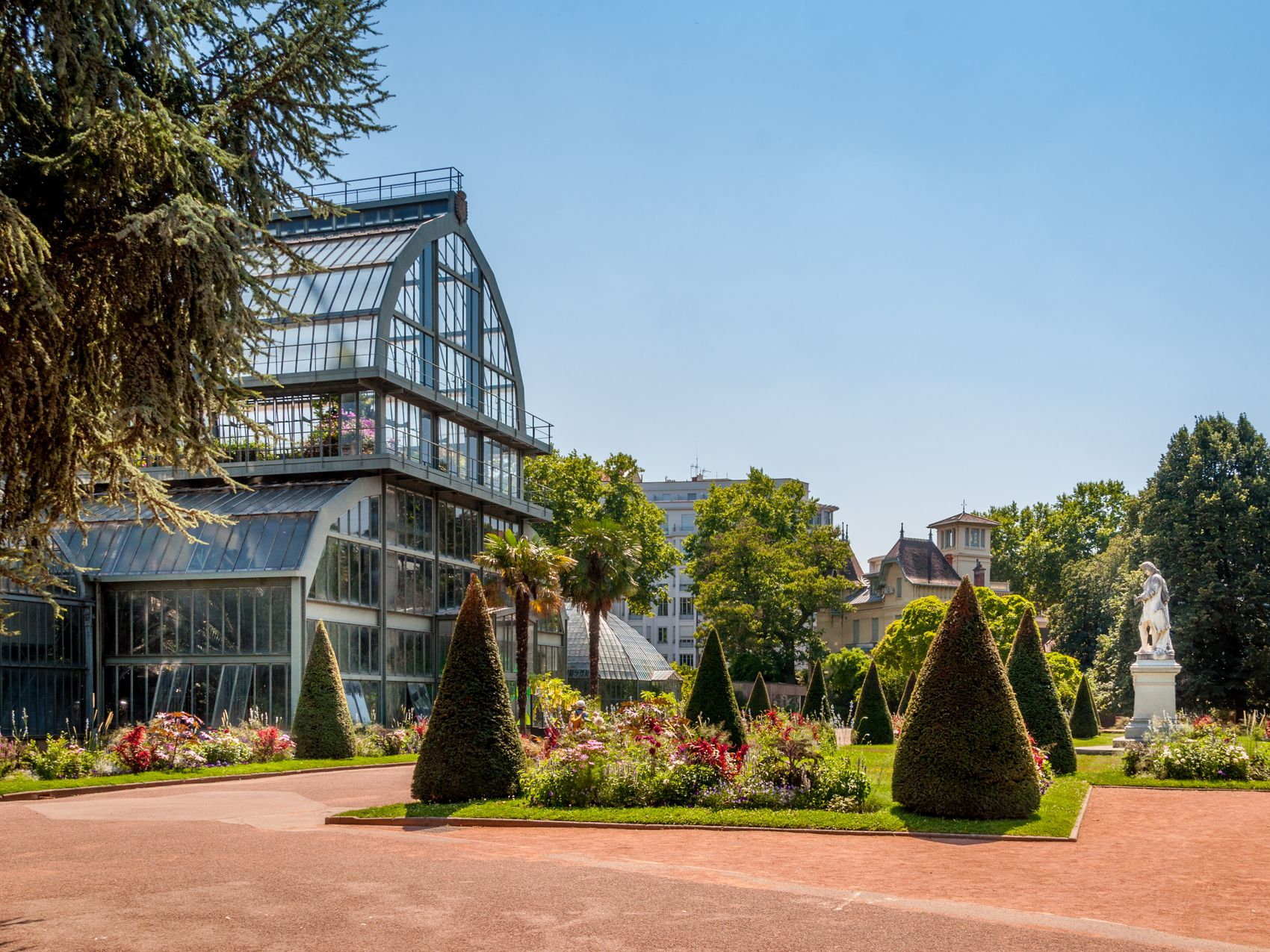 Une véranda en verre à structure métallique se dresse dans un jardin public ensoleillé.