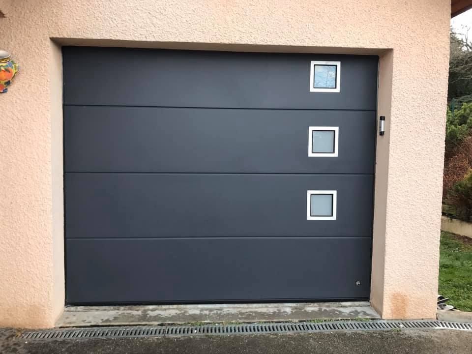 Porte de garage sectionnelle moderne gris foncé avec trois fenêtres carrées alignées verticalement sur le côté droit.