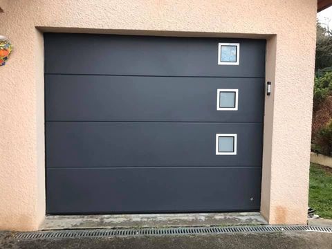 Porte de garage sectionnelle moderne gris foncé avec trois fenêtres carrées alignées verticalement sur le côté droit.