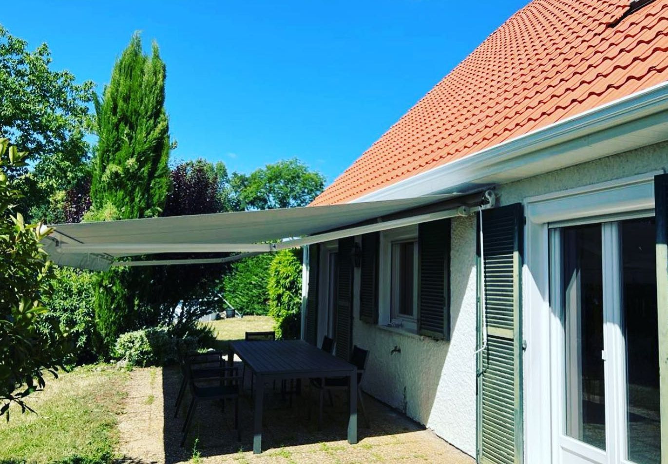 Une terrasse ombragée par un auvent rétractable gris clair fixé sur le côté d'une maison blanche.