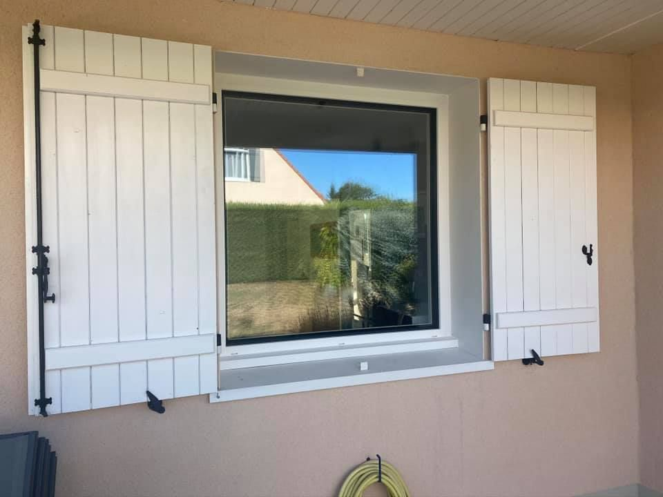 Un mur extérieur blanc, percé d'une fenêtre aux volets en bois blancs ouverts.