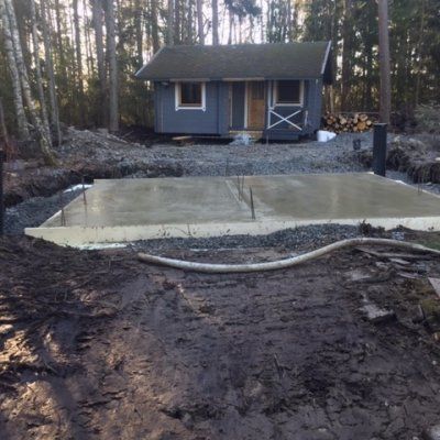 Tuorevalettu betonilaatta seisoo mutaisella, metsäisellä alueella, ja taustalla näkyy pieni harmaa mökki.