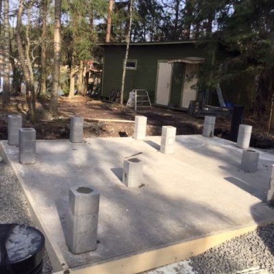 Betonilaatan perustus, jossa on useita pystyssä olevia harkkopilareita metsäisellä pihalla, taustalla pieni vaja.