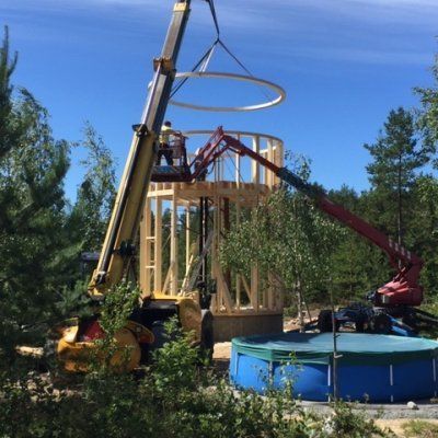 Rakennusnosturi nostaa metallirenkaan pyöreän rakennelman puurunkoon sinisen uima-altaan viereen.