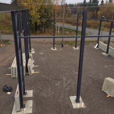Rakennustyömaa, jossa näkyy teräksisiä tukipilareita ja betonisia antureita soratolla lähellä tietä ja metsää.