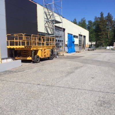Keltainen saksinostin pysäköitynä suuren teollisuusrakennuksen viereen, jonka sinisen pressuoven edessä on rakennustelineet.