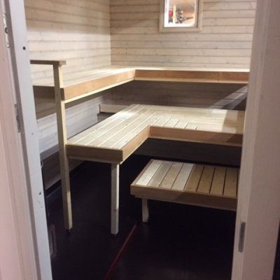 Sisäkuva saunasta, jossa on vaaleat puupaneelit, L-kirjaimen muotoiset porrastetut lauteet ja pieni askelmajakkara tummalla lattialla.