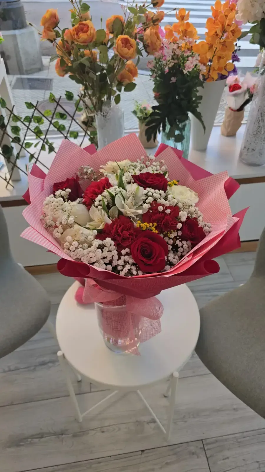 Ramo de flores rojas y blancas envuelto en papel rosa sobre una pequeña mesa blanca, con flores de fondo borrosas.