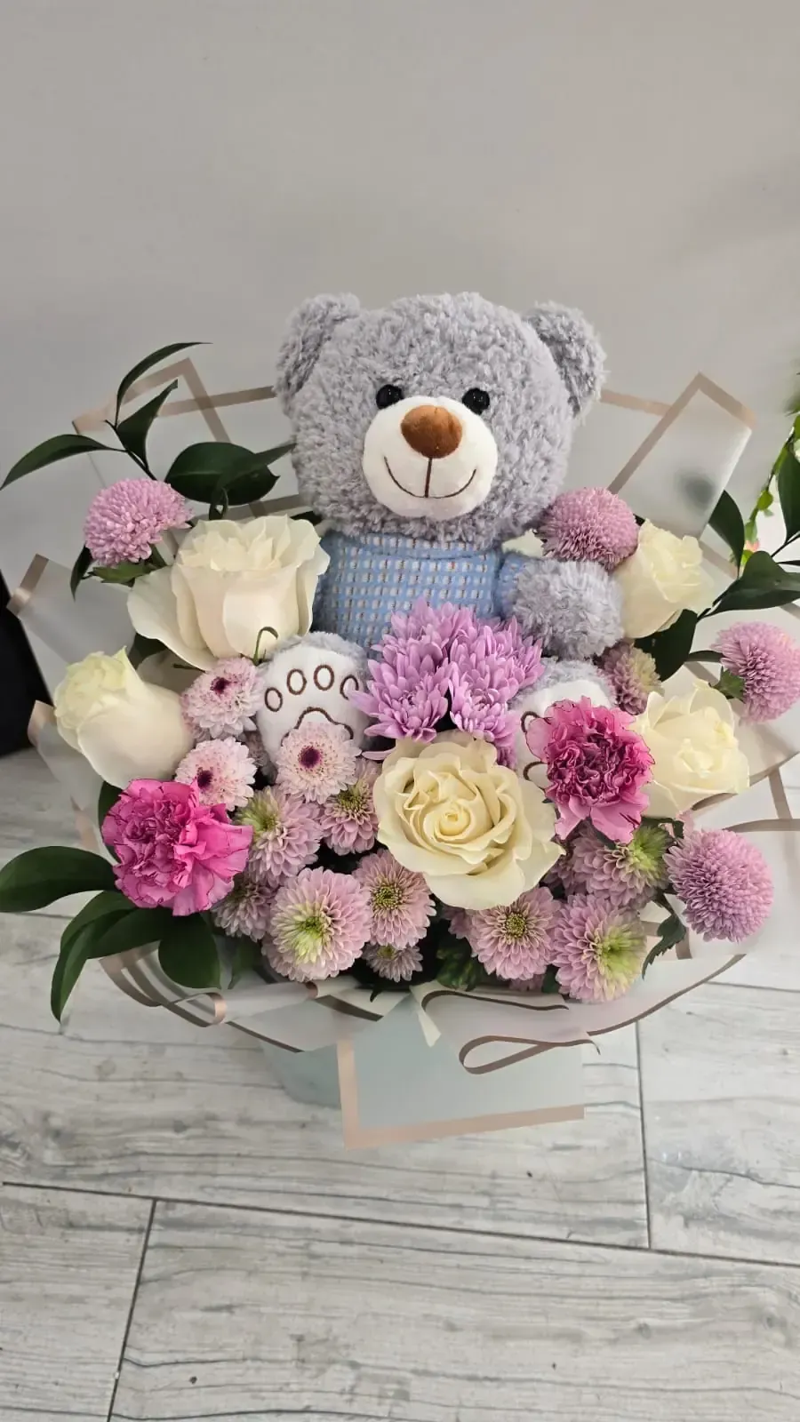Osito de peluche acurrucado en un ramo de flores de rosas blancas, claveles rosados y crisantemos morados.