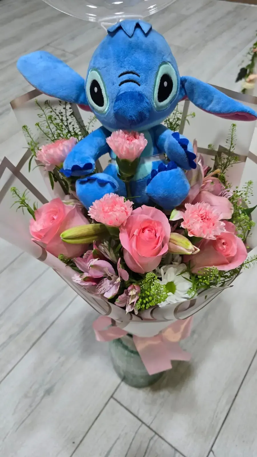 Peluche de Stitch sentado en un ramo floral con rosas rosas y claveles, envuelto en papel.