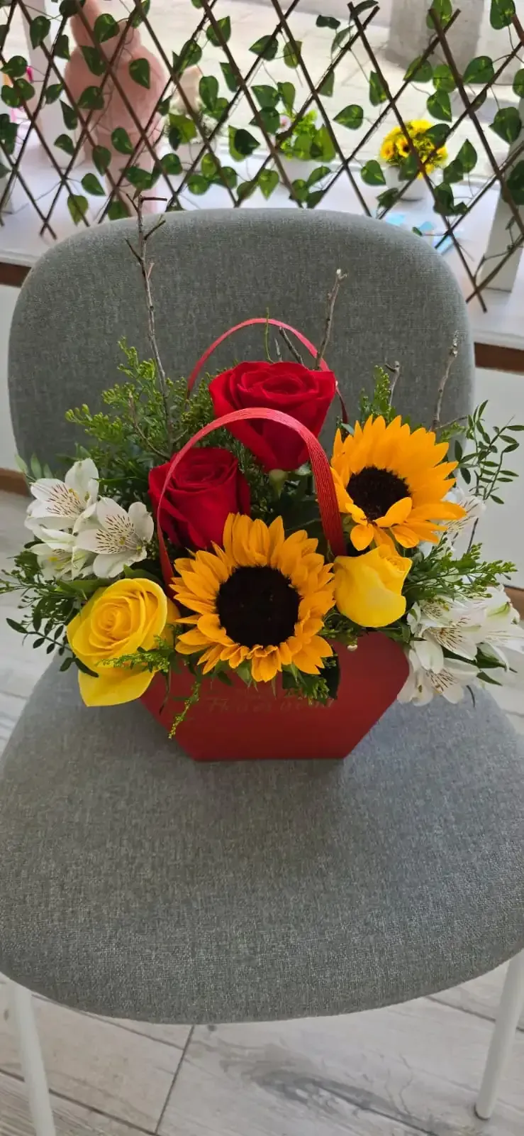 Una canasta de flores rojas con girasoles, rosas rojas y rosas amarillas sobre una silla gris.