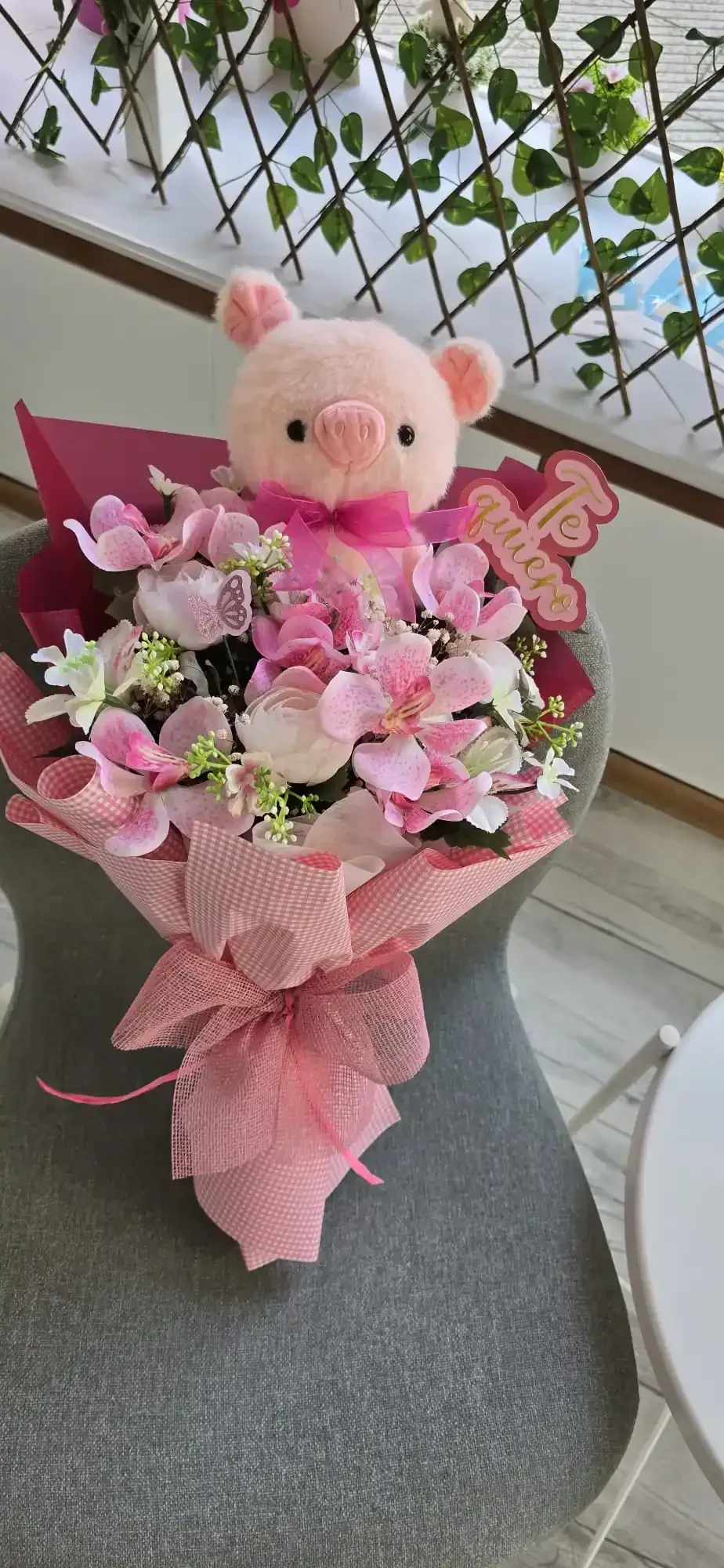Ramo de flores rosas, peluche de cerdo y lazo rosa sobre silla gris.
