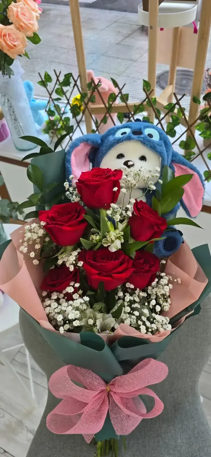 Ramo de rosas rojas, velo de novia y peluche de Stitch, envuelto en papel rosa y verde azulado con un lazo rosa.