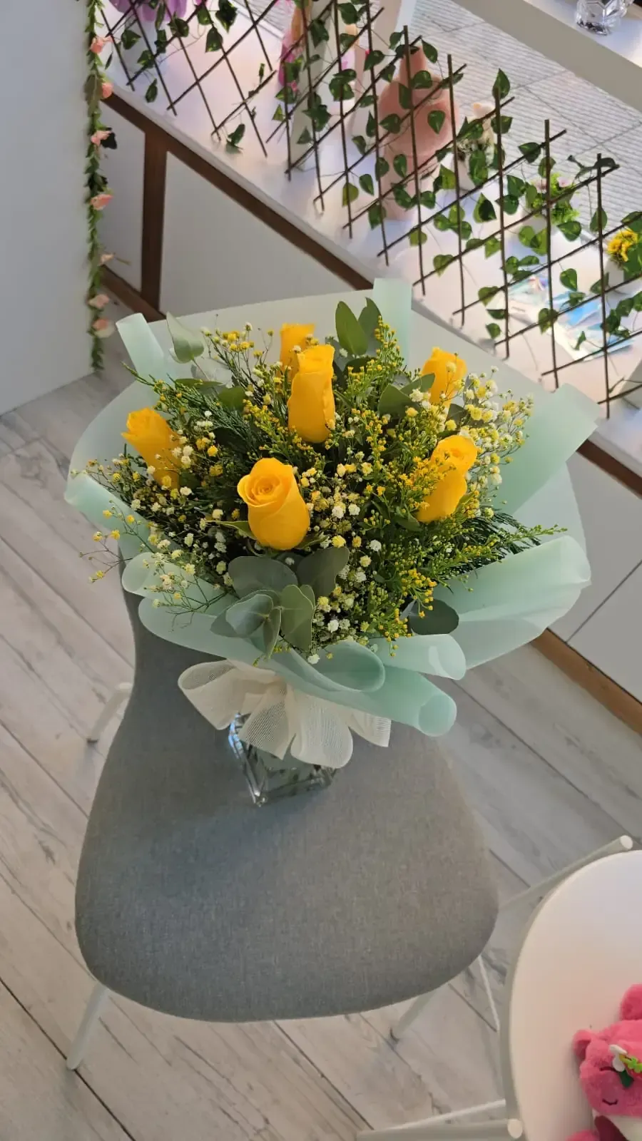 Ramo de rosas amarillas, follaje y gitanos, envuelto en papel verde claro y atado con una cinta blanca, sobre una silla gris.