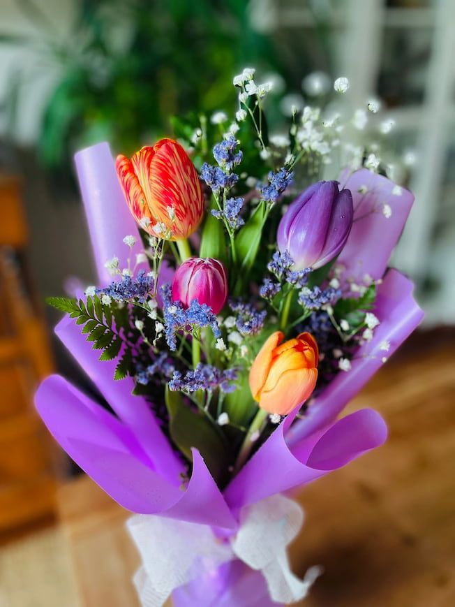 Ramo de tulipanes de colores y gitanos envuelto en papel morado, atado con una cinta blanca.